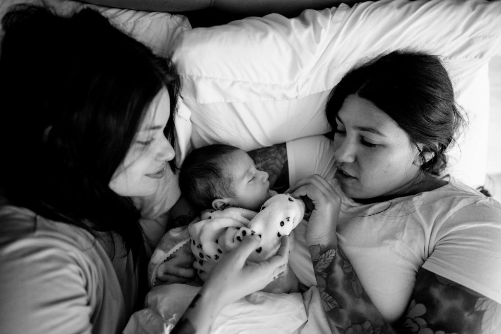 Female couple embracing baby in bed.